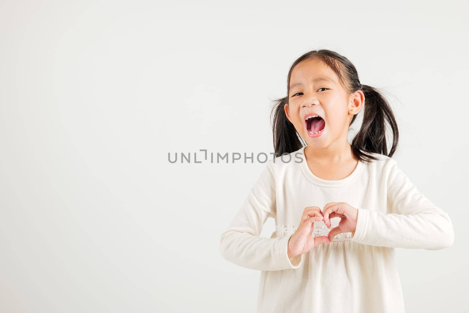 A smiling Asian young kid girl shows confidence, creating a heart shape with fingers and hands on white background. Portrait of kindergarten child sending love for Valentine's Day