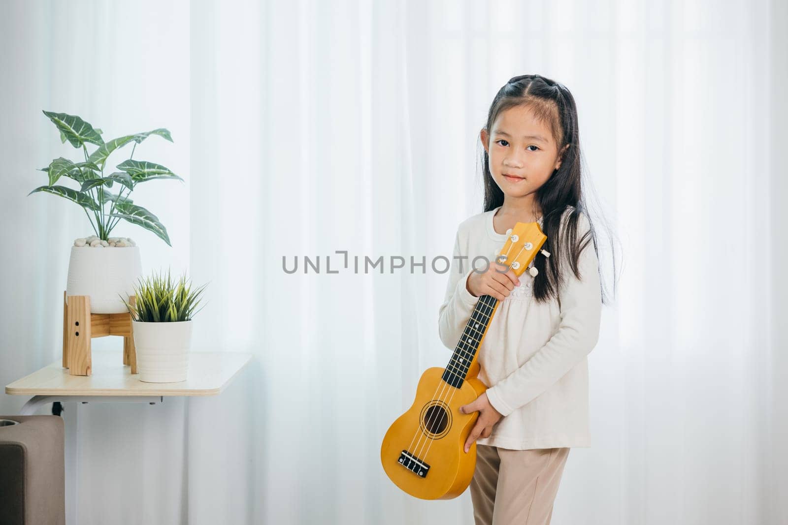 Asian girl strums a ukulele while standing in a cozy, well-decorated living room. Her cheerful expression and love for music harmonize perfectly with the modern, softly lit environment.