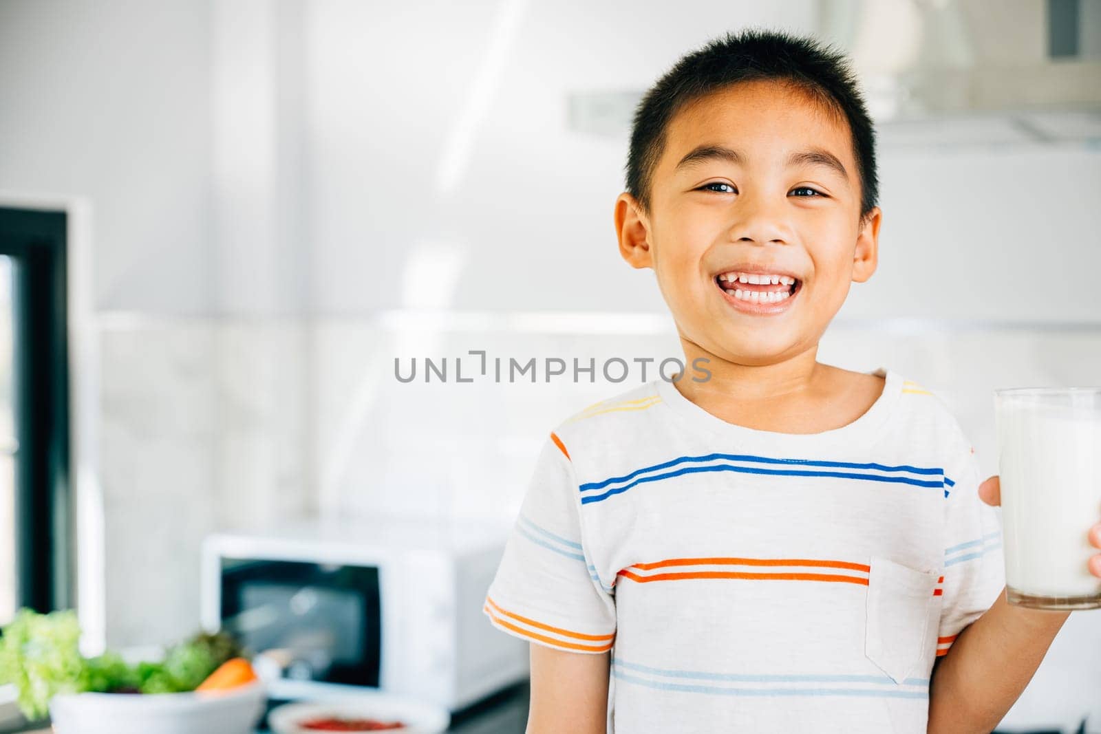Cheerful Asian little boy holds milk in kitchen, smiling happily. Portrait of cute son enjoying drink. Joyful child sips calcium-rich liquid, radiating happiness at home give me.