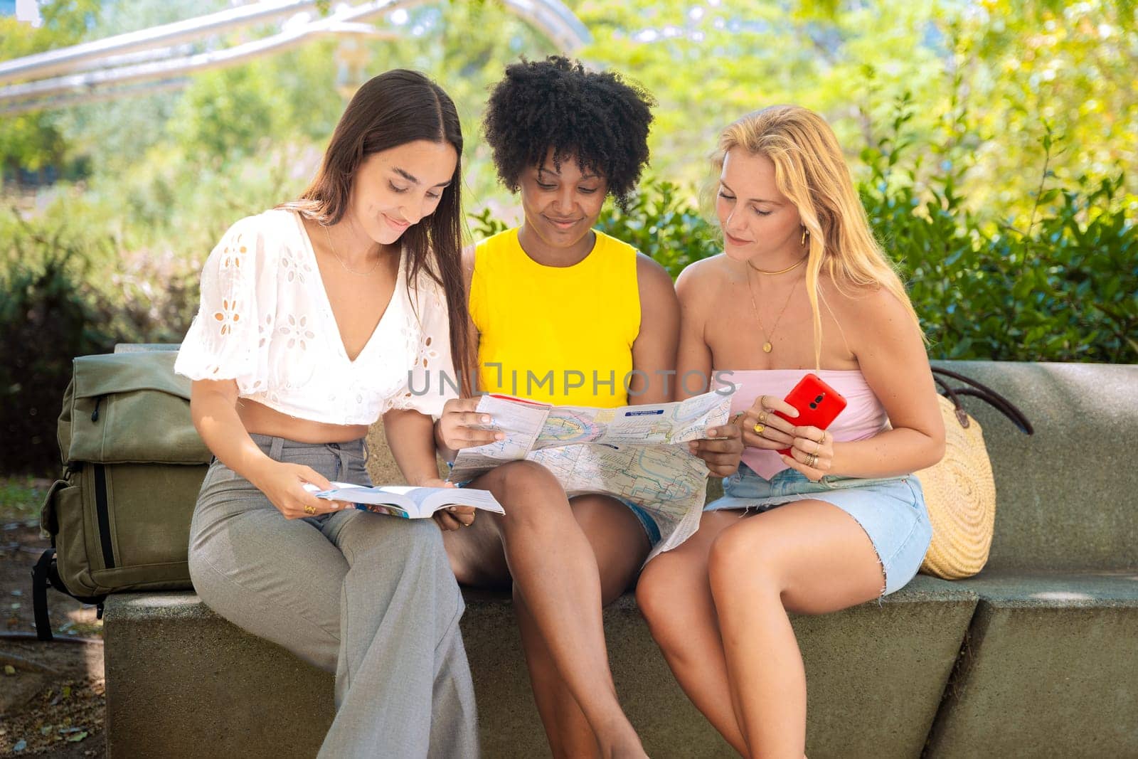 Three happy young women stand outdoors, looking at maps and a travel guide. They smile while planning their trip, enjoying a fun adventure in a sunny urban setting with trees and buildings.
