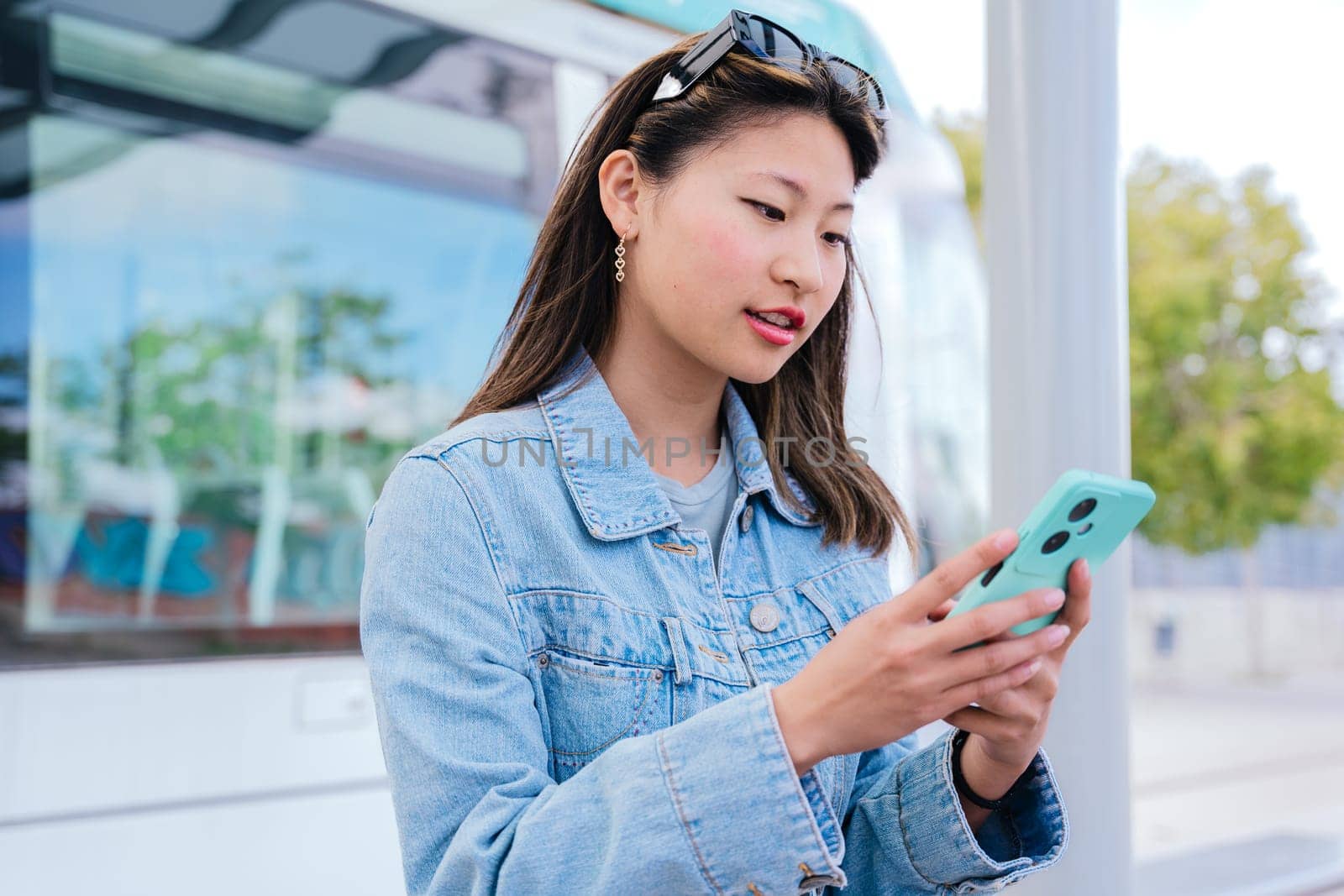 A stylish young woman in a denim jacket checks her smartphone while waiting at a tram station. She appears focused and relaxed, with sunglasses on her head. A modern urban lifestyle scene.