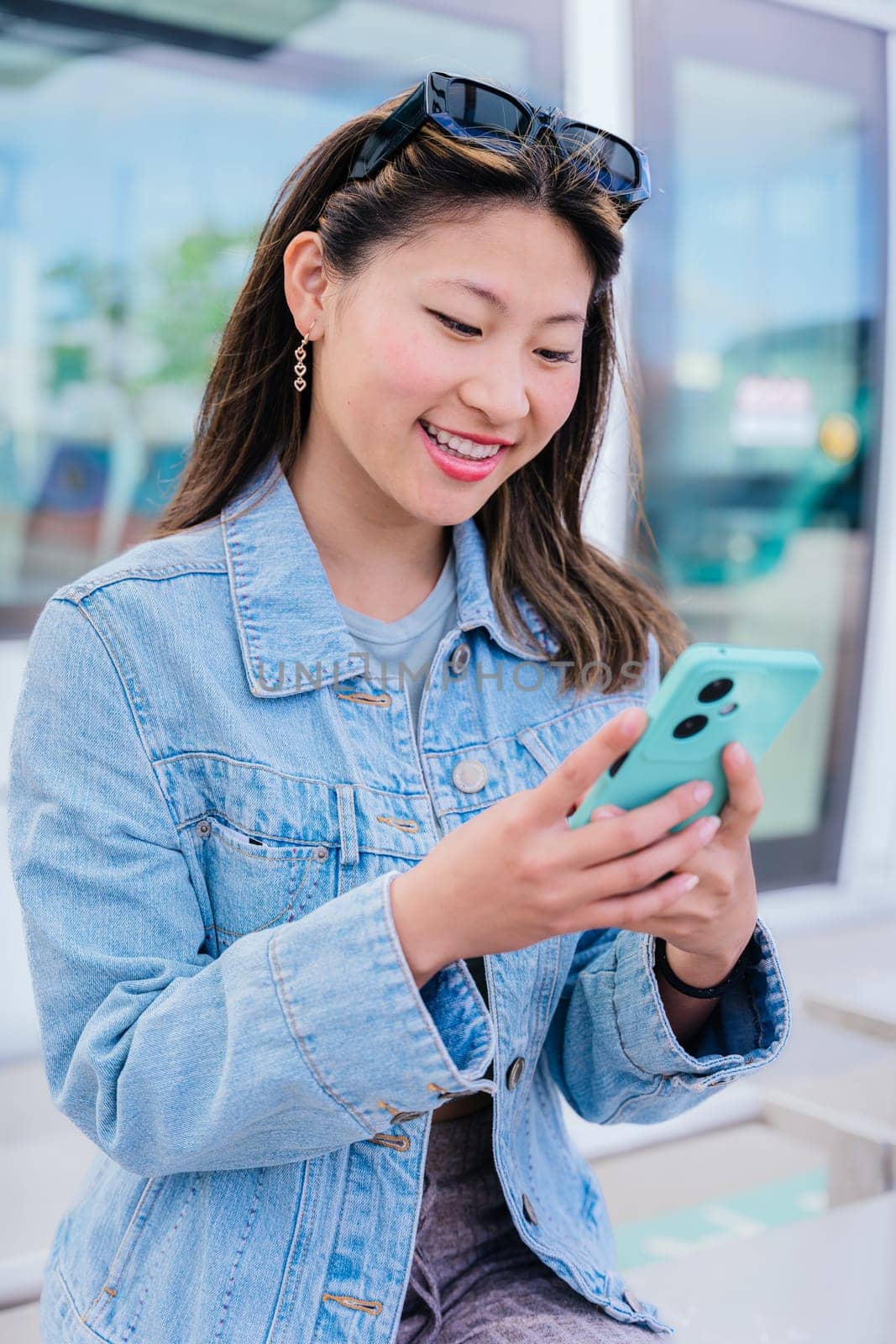 A stylish young woman in a denim jacket checks her smartphone while waiting at a tram station. She appears focused and relaxed, with sunglasses on her head. A modern urban lifestyle scene.