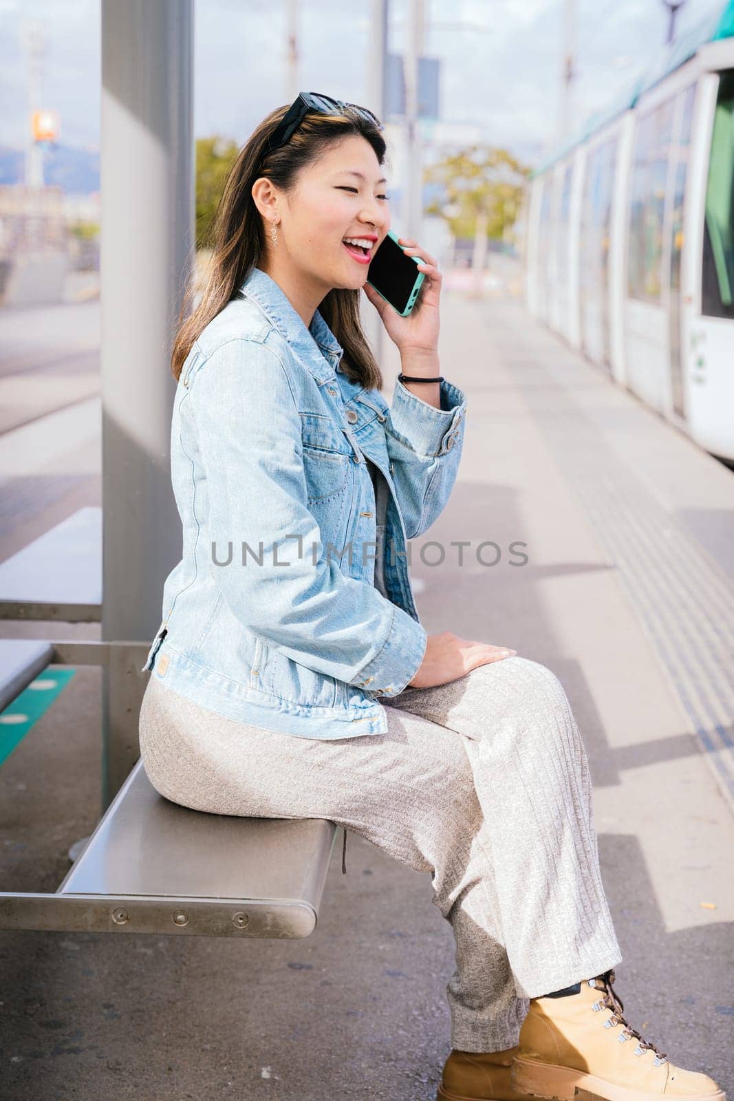 Smiling young woman wearing casual clothes and denim jacket sitting at a tram stop, talking on her mobile phone. Urban setting with tram and city background. Bright and sunny day.