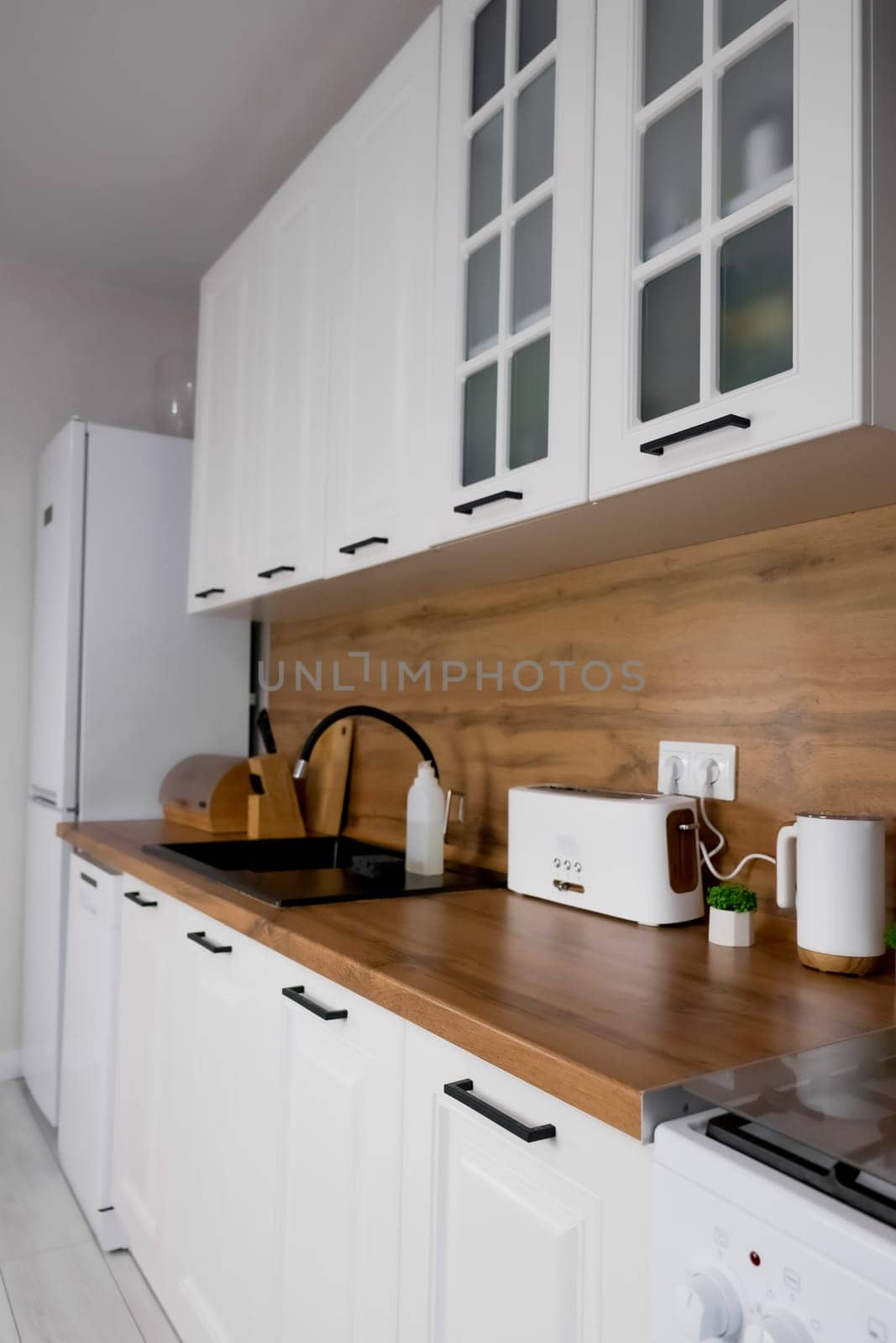 Horizontal photo, side view of a modern kitchen with a white cabinet and a wooden countertop on which there is a toaster, plants and a bread box with a knife rack, a prominent gas stove, a black sink and a white refrigerator. Modern kitchen concept, kitchen island, modern kitchen style. High quality photo