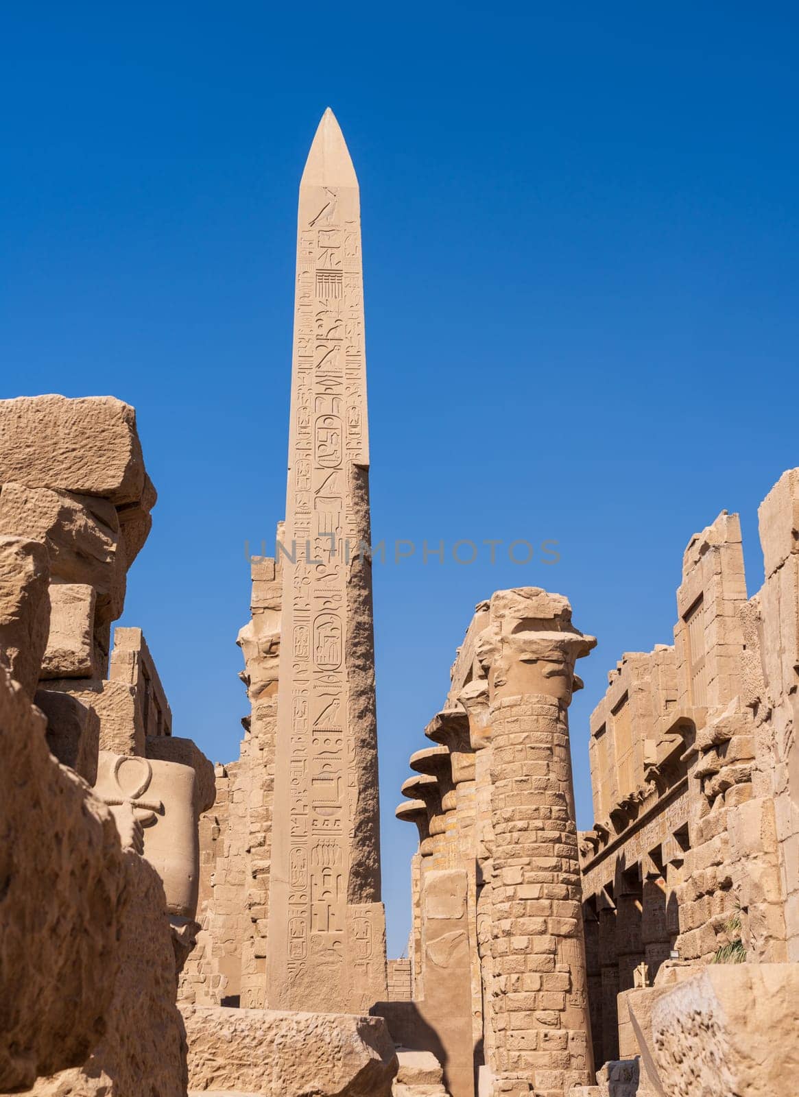 Ancient Egyptian obelisk covered in hieroglyphs stands tall against a clear blue sky, surrounded by ruins of Karnak temple