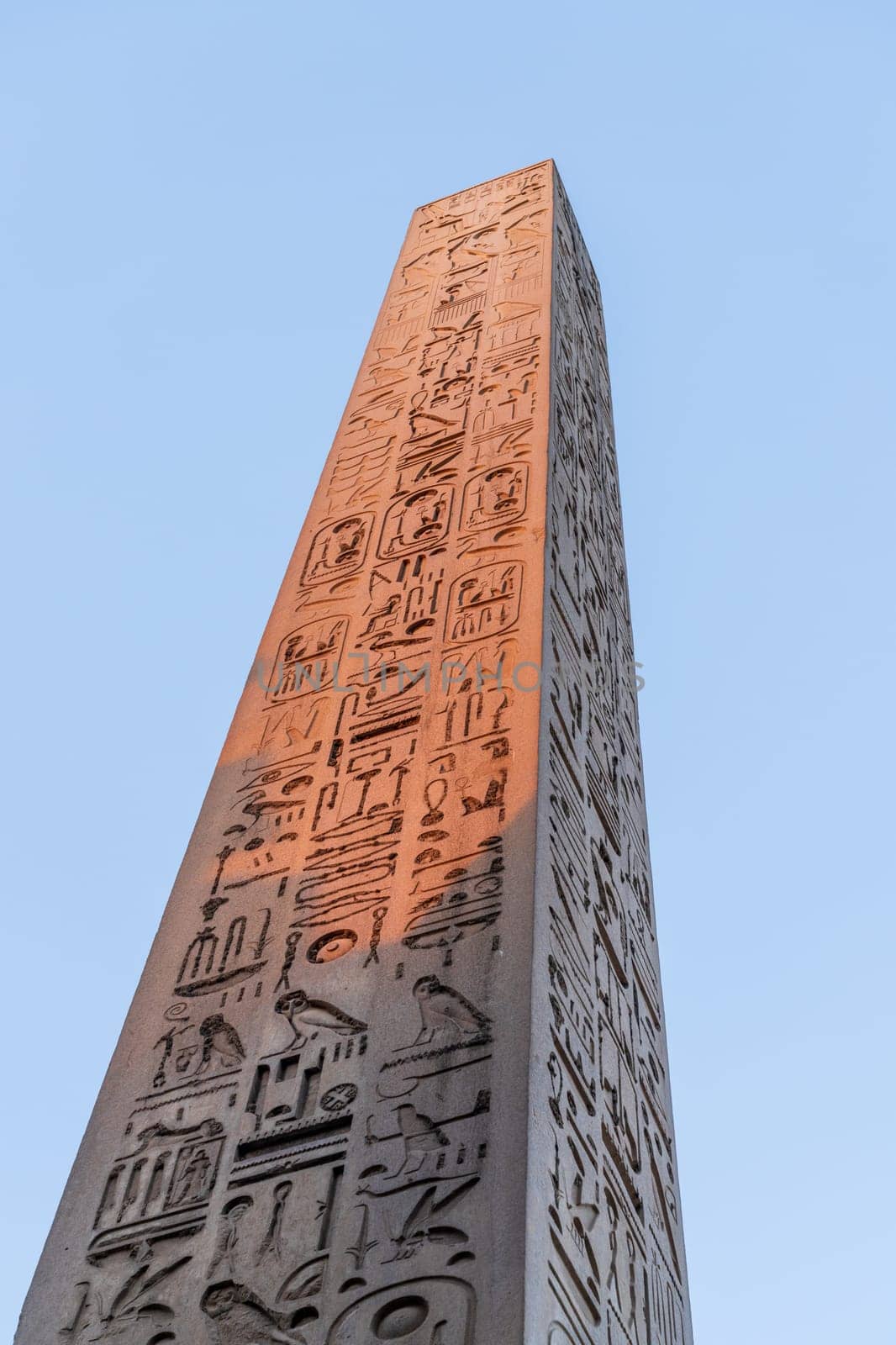 Ancient Egyptian obelisk standing tall against a clear blue sky, its surface covered in intricate hieroglyphs. Placed at the entrance of Luxor temple