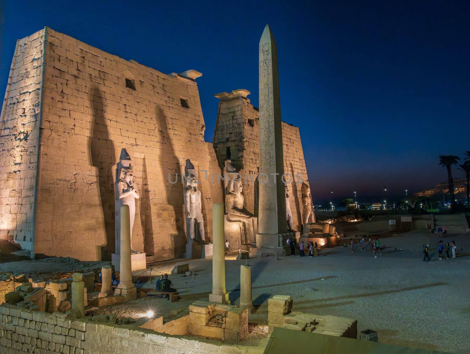 Massive stone wall and standing and seated statues with obelisk in Luxor Temple in Egypt illuminated at night