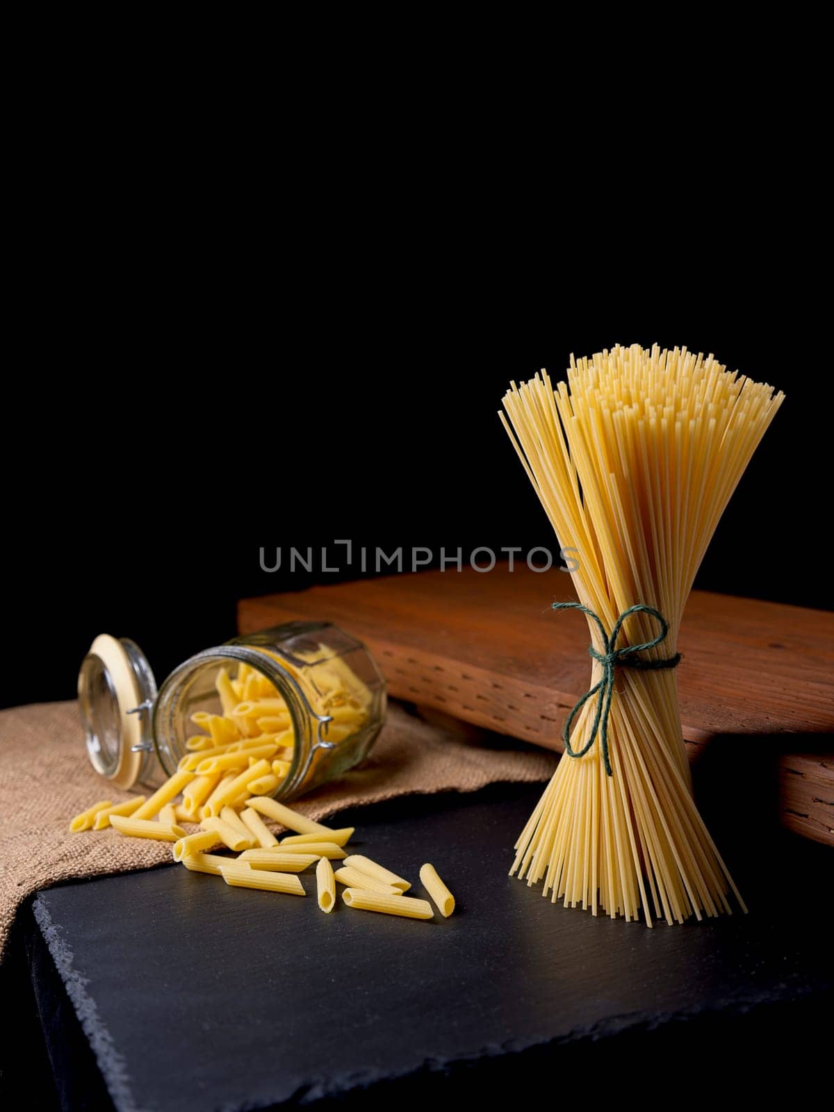 A still life photo of dried spaghetti noodles standing up and penne noodles spilling out of a glass jar.