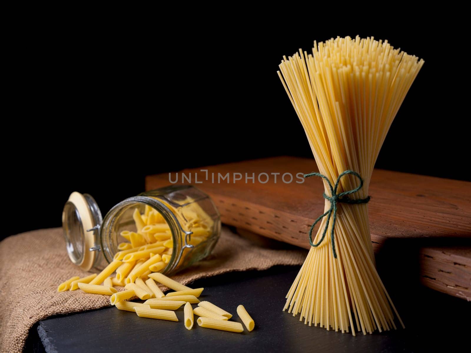 A still life photo of dried spaghetti noodles standing up and penne noodles spilling out of a glass jar.