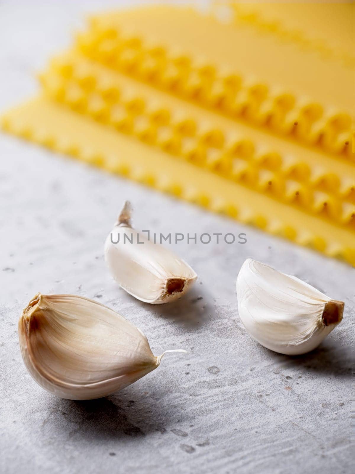 A closeup studio photo of three cloves of garlic and dried lasagna noodle strips.