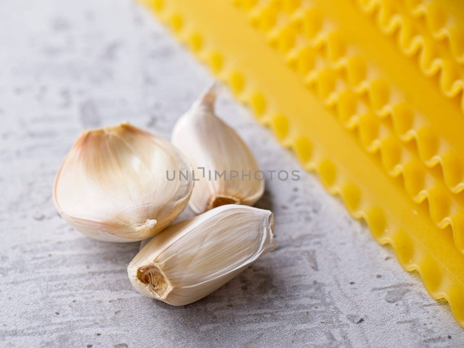 A closeup studio photo of three cloves of garlic and dried lasagna noodle strips.