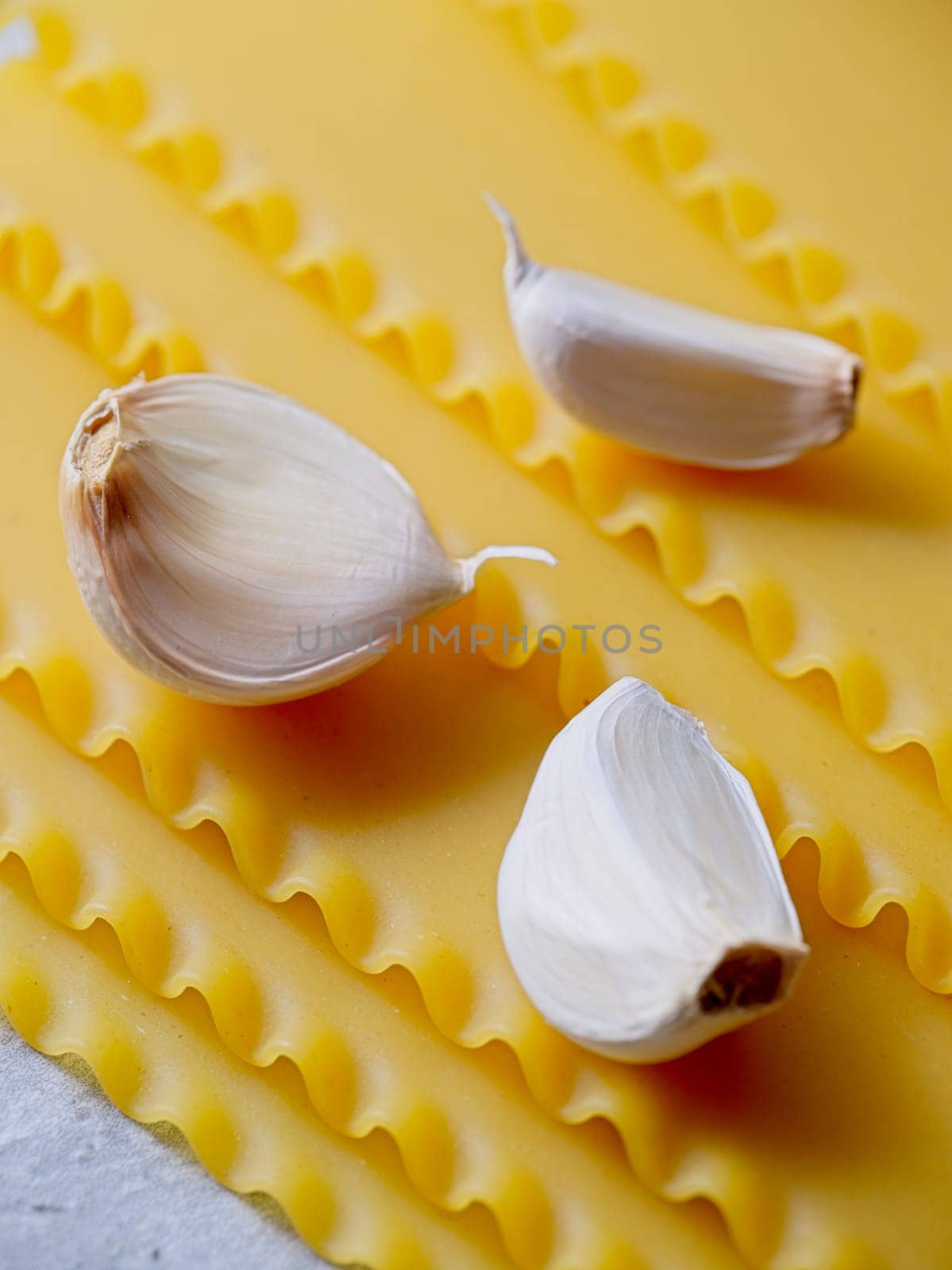 A closeup studio photo of three cloves of garlic and dried lasagna noodle strips.