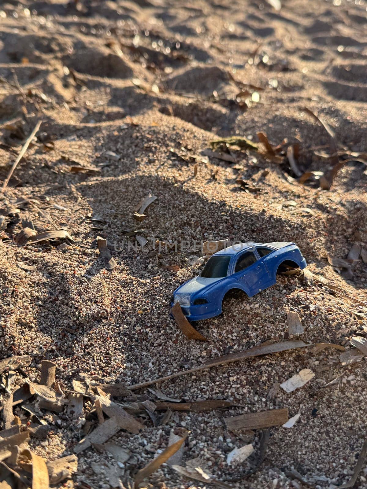 A blue toy car partially buried in sand on a beach, surrounded by textured sand and small twigs under warm sunlight.