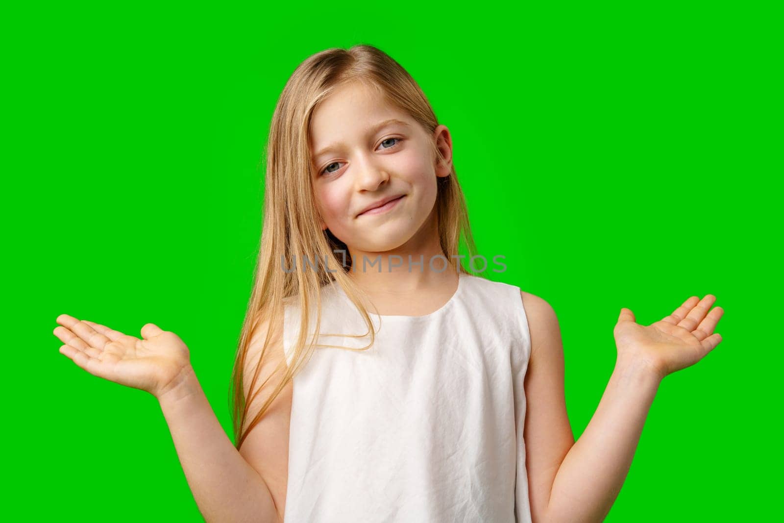 A young girl with long hair smiles and shrugs while standing against a bright green background in studio