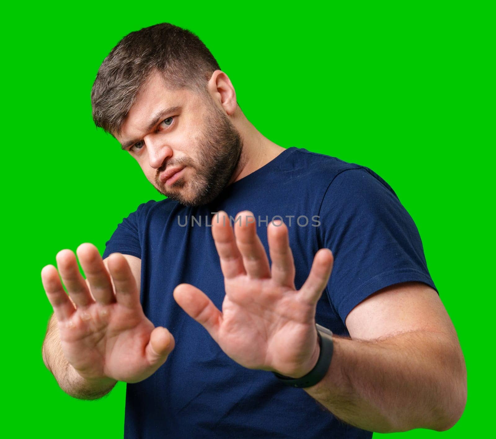 A man with a serious expression holds up both hands in a stopping gesture against a bright green background in studio