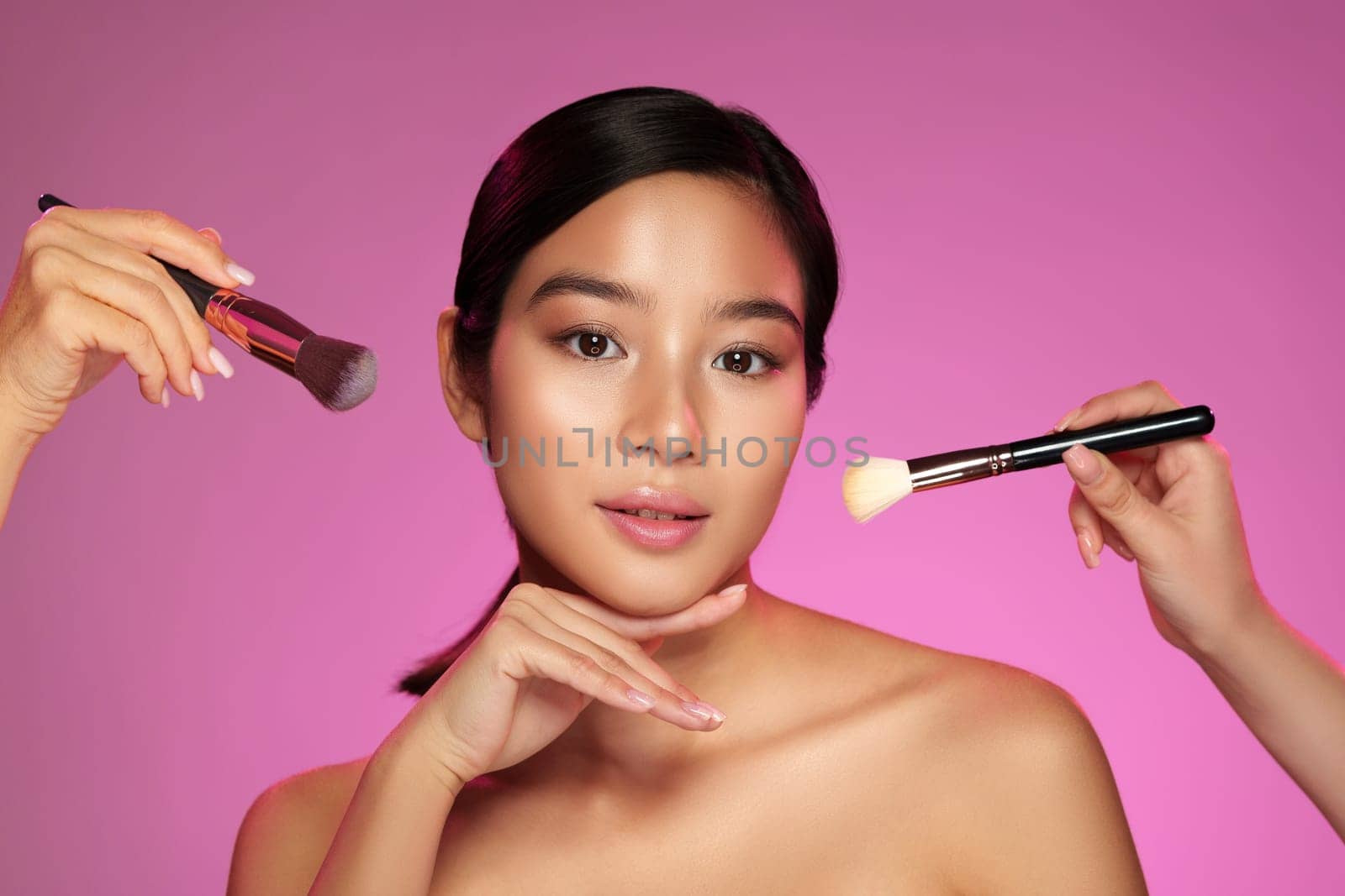 A model poses with a serene expression as makeup artists apply products using brushes. The bright pink background enhances the beauty-focused atmosphere and highlights the artistry involved.