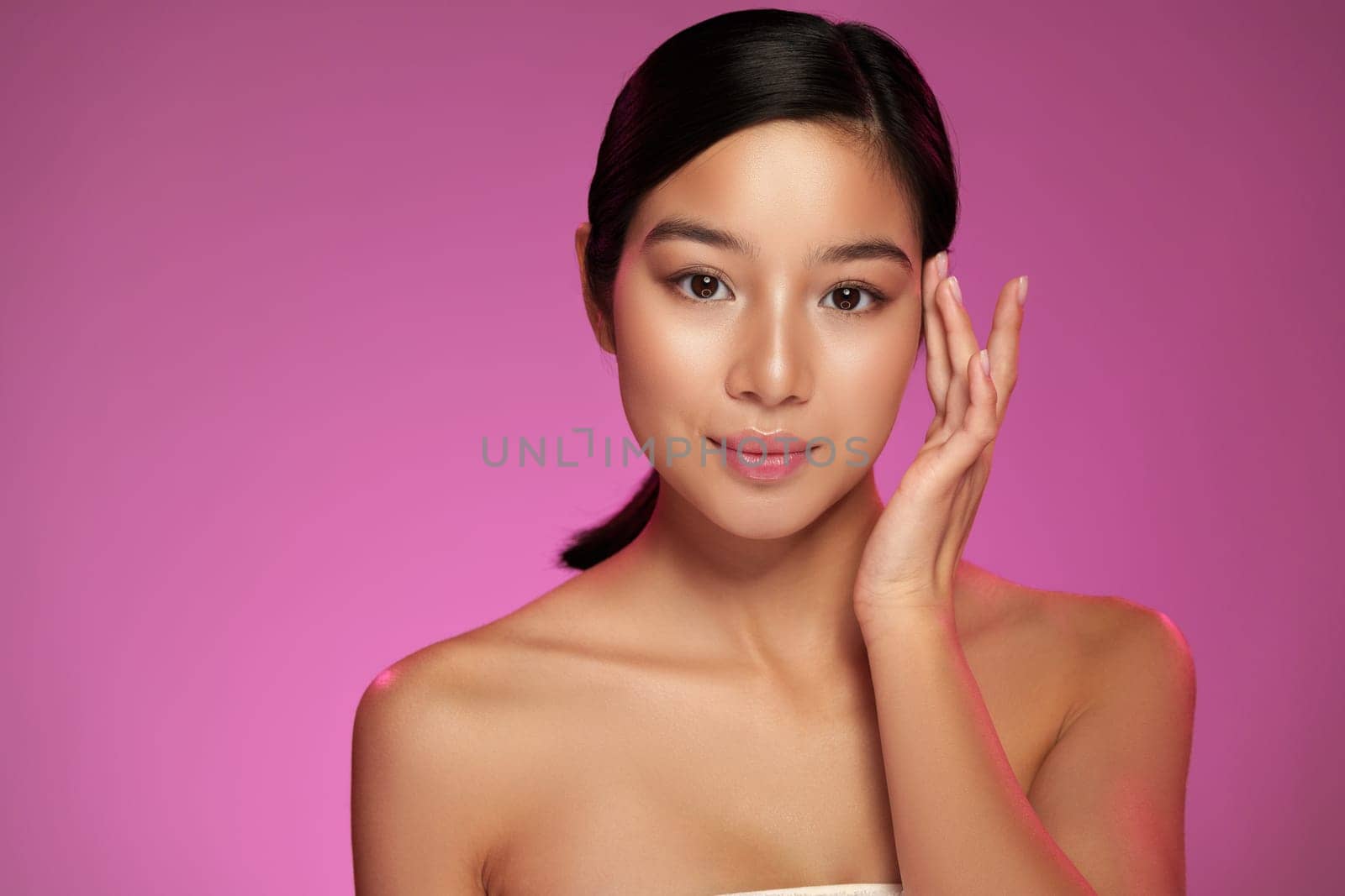 A woman showcases her flawless complexion while gently touching her face. She poses confidently against a vibrant pink backdrop, emphasizing beauty and skincare.