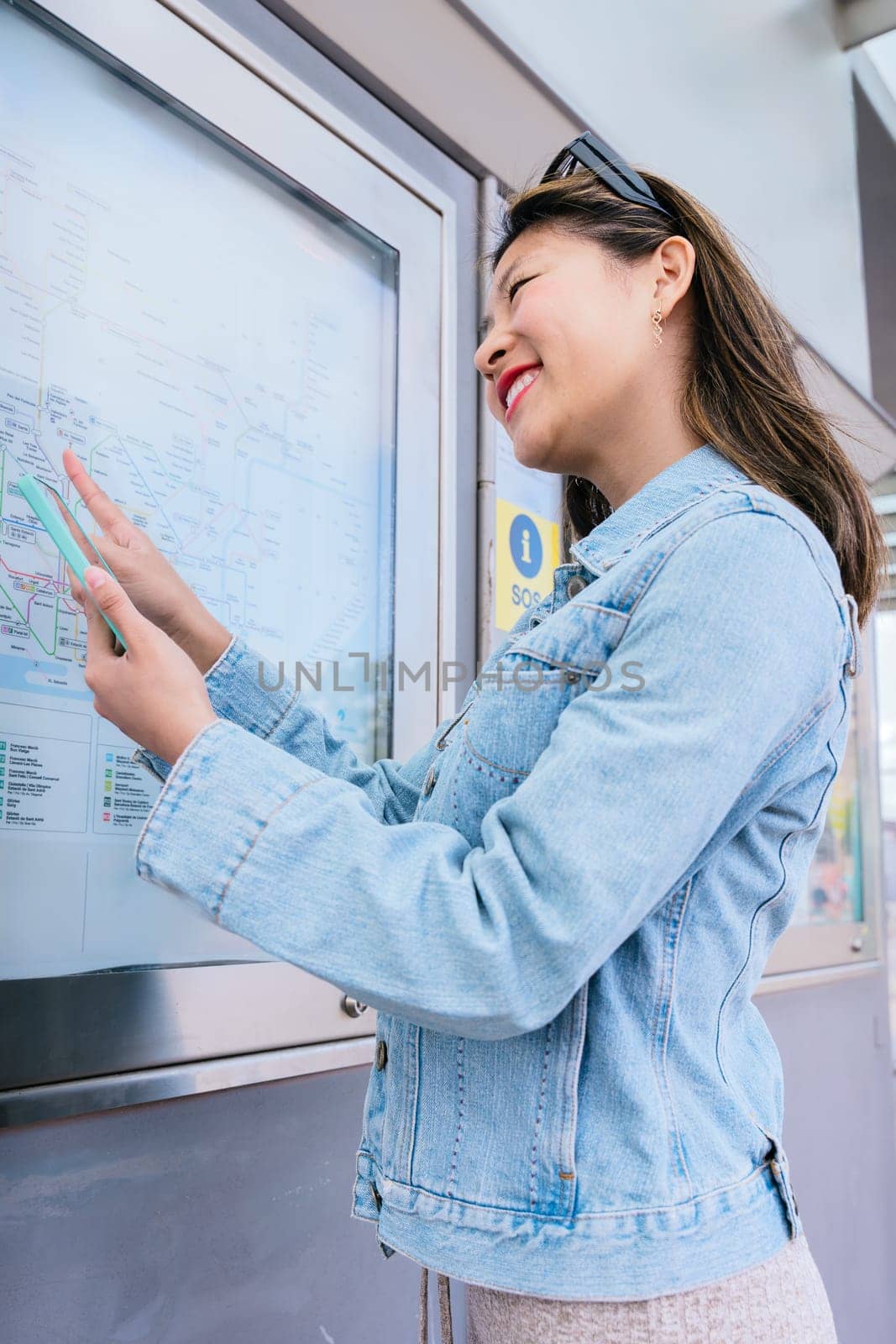 Cheerful young woman in a denim jacket checking a public transport map while holding her smartphone. She smiles as she plans her route at a tram stop in an urban setting.