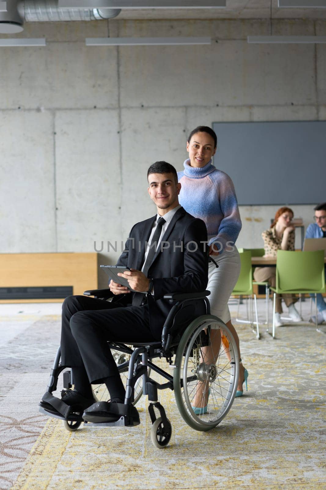 In a professional office setting, a businesswoman helping her director, who is sitting in a wheelchair and using a tablet, while their colleagues collaborate in the background