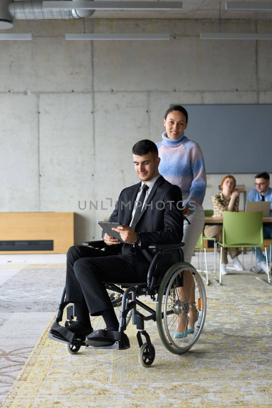 In a professional office setting, a businesswoman helping her director, who is sitting in a wheelchair and using a tablet, while their colleagues collaborate in the background