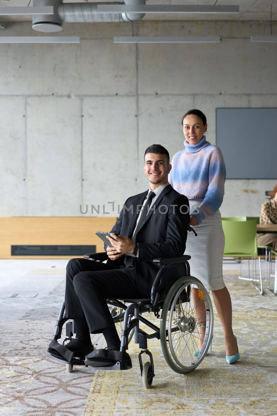 In a professional office setting, a businesswoman helping her director, who is sitting in a wheelchair and using a tablet, while their colleagues collaborate in the background