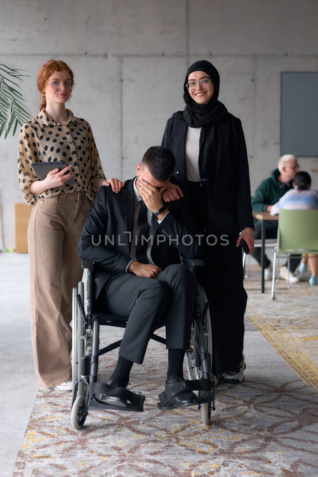 A diverse business team poses confidently, including a smiling director seated in a wheelchair, flanked by two professional female colleagues, one wearing a hijab and the other holding a tablet, in a modern office environment.