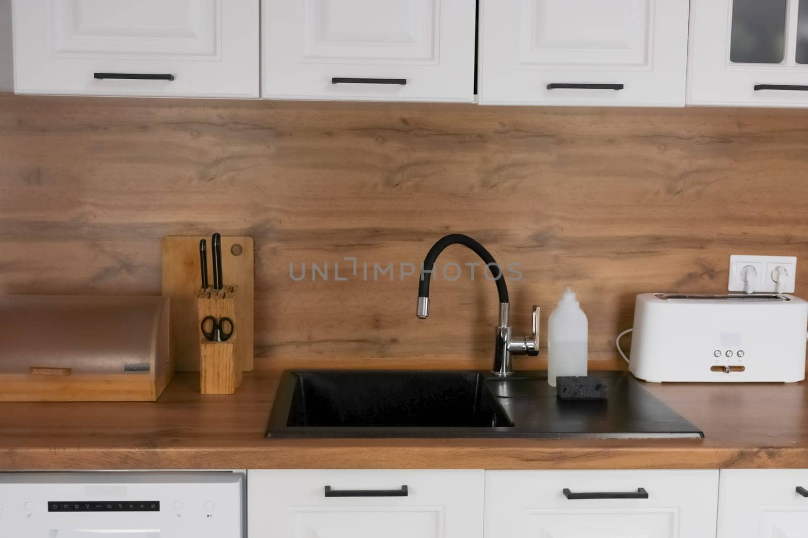A black kitchen sink with a black flexible faucet, it has dishwashing detergent in a white bottle and a black dishwashing sponge against the background of a modern kitchen with white cabinets and a wooden countertop, there are kitchen utensils on the table. Dishwashing concept, kitchen island, modern interiors. High quality photo