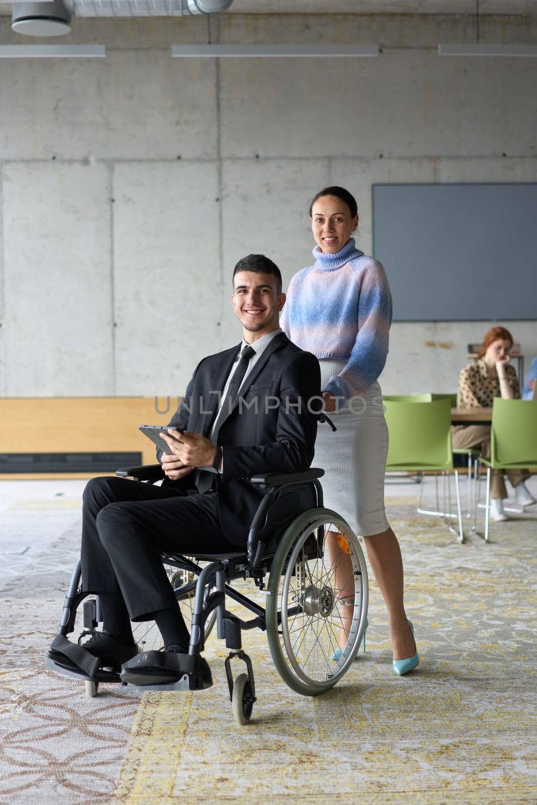 In a professional office setting, a businesswoman helping her director, who is sitting in a wheelchair and using a tablet, while their colleagues collaborate in the background