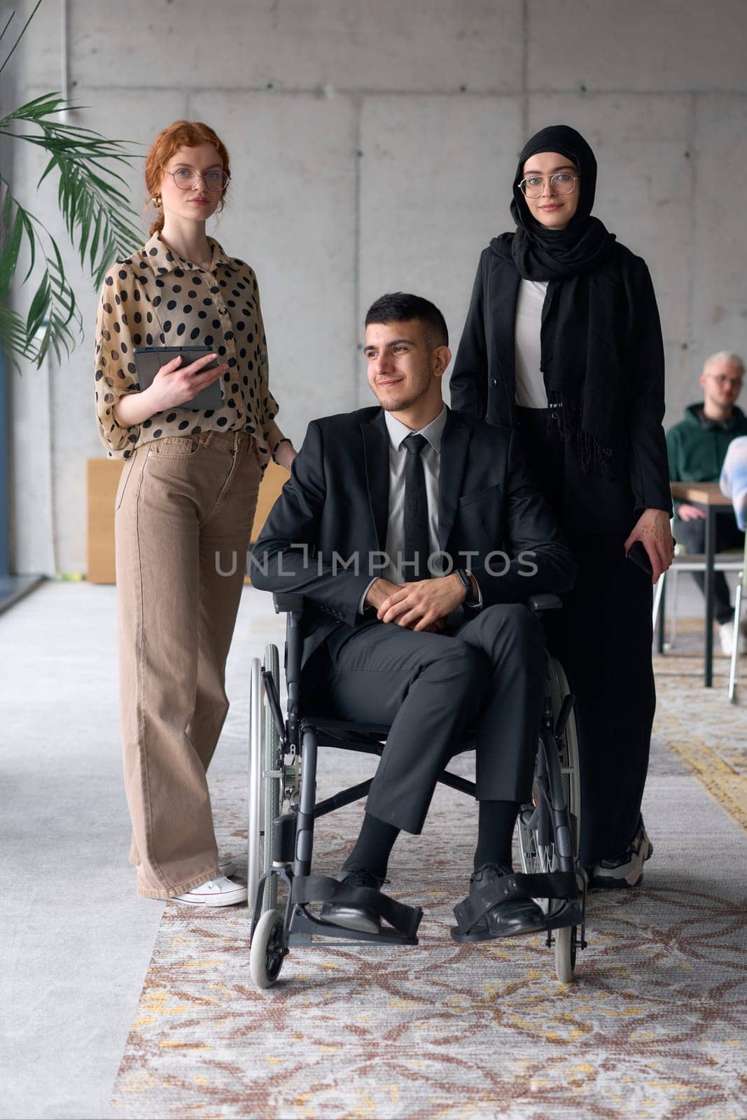 A diverse business team poses confidently, including a smiling director seated in a wheelchair, flanked by two professional female colleagues, one wearing a hijab and the other holding a tablet, in a modern office environment.