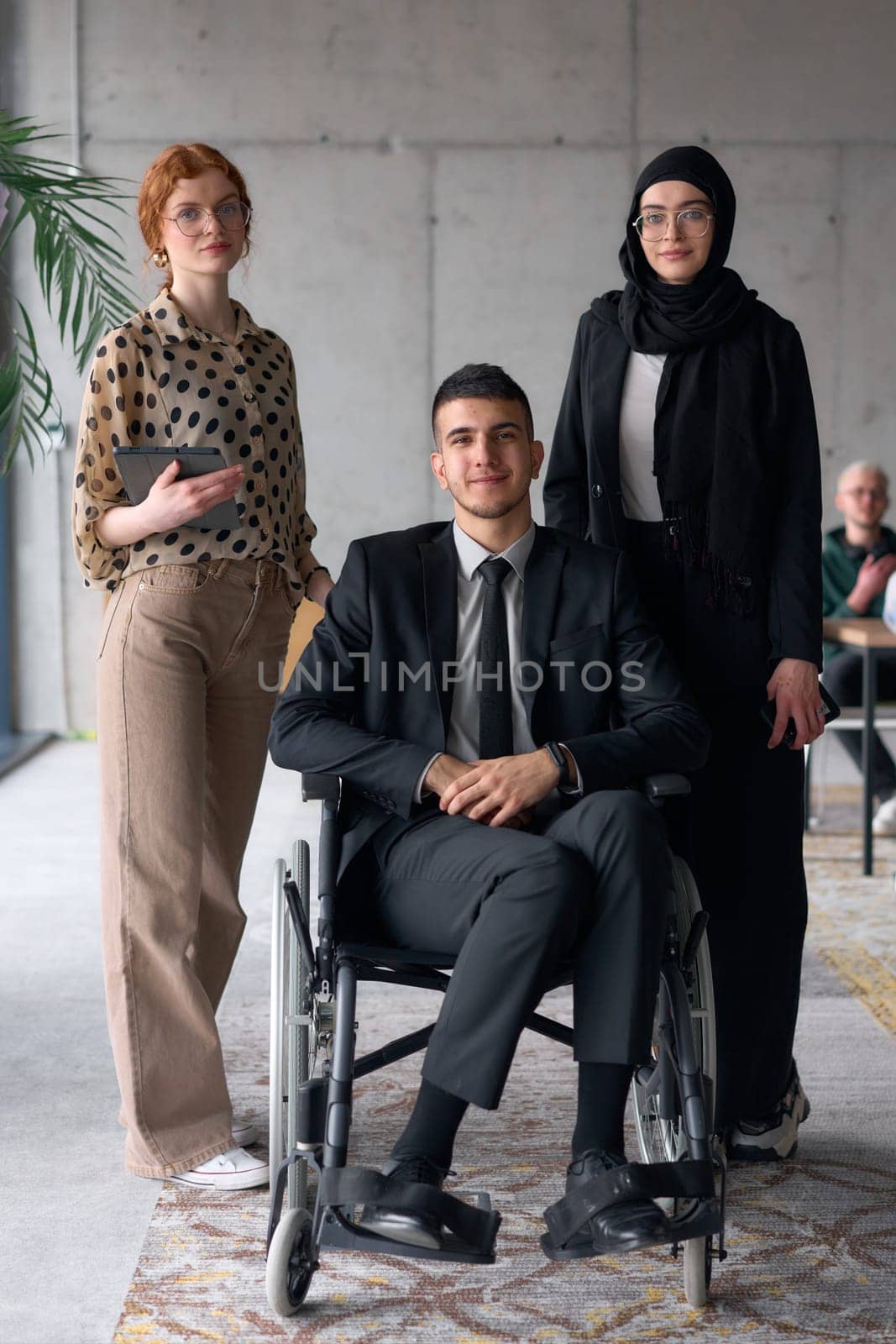 A diverse business team poses confidently, including a smiling director seated in a wheelchair, flanked by two professional female colleagues, one wearing a hijab and the other holding a tablet, in a modern office environment.