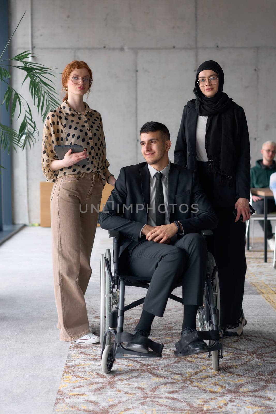 A diverse business team poses confidently, including a smiling director seated in a wheelchair, flanked by two professional female colleagues, one wearing a hijab and the other holding a tablet, in a modern office environment.