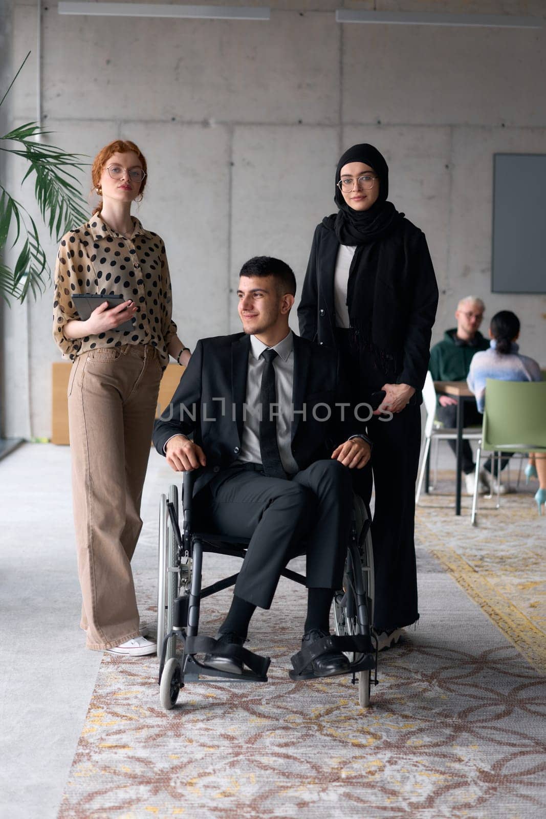 A diverse business team poses confidently, including a smiling director seated in a wheelchair, flanked by two professional female colleagues, one wearing a hijab and the other holding a tablet, in a modern office environment.