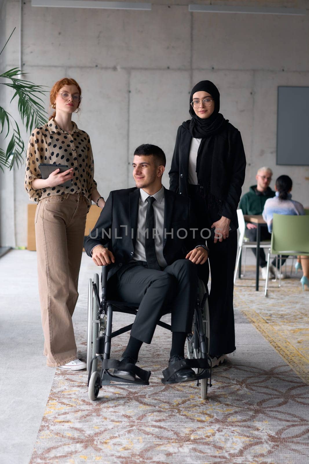 A diverse business team poses confidently, including a smiling director seated in a wheelchair, flanked by two professional female colleagues, one wearing a hijab and the other holding a tablet, in a modern office environment.