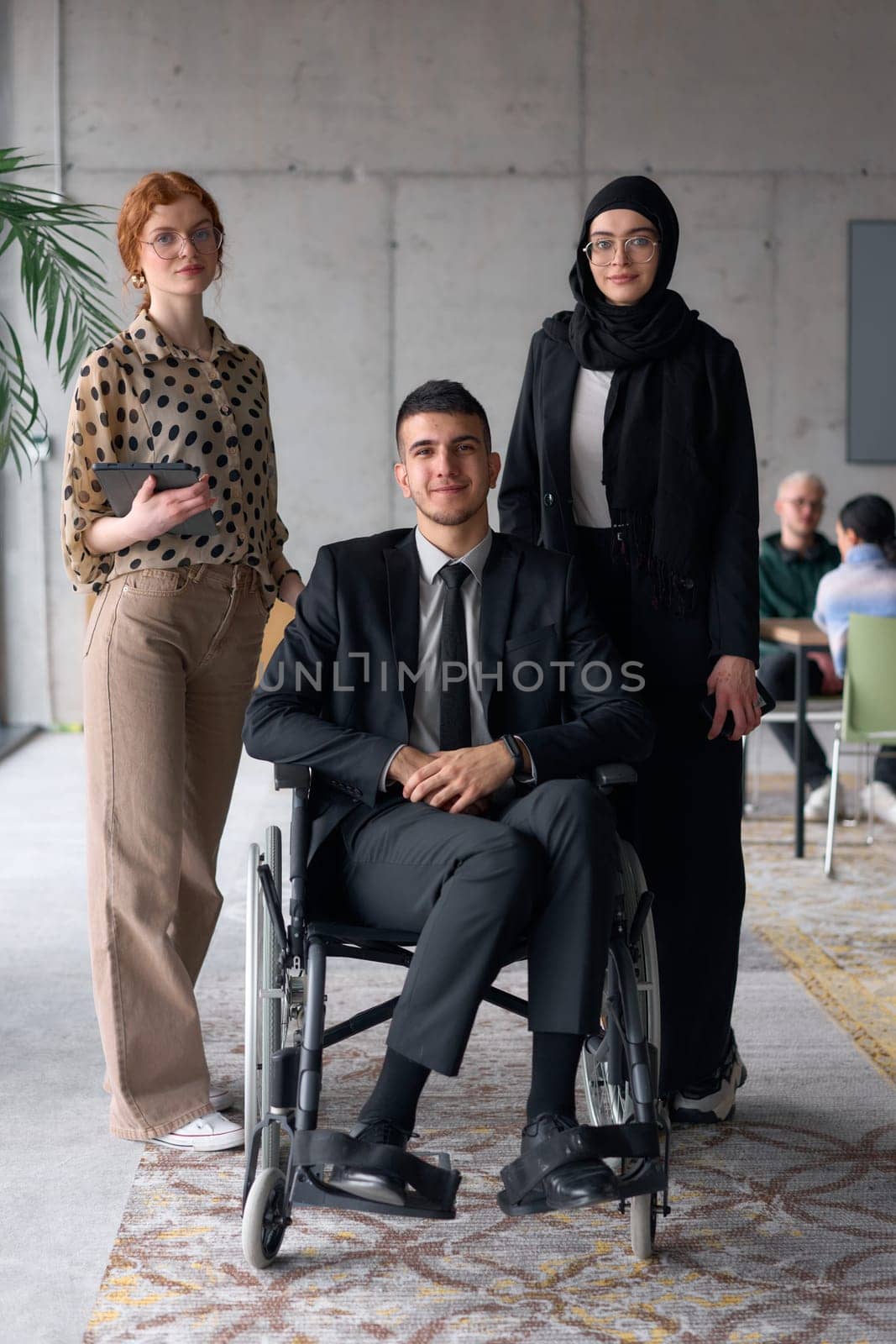 A diverse business team poses confidently, including a smiling director seated in a wheelchair, flanked by two professional female colleagues, one wearing a hijab and the other holding a tablet, in a modern office environment.