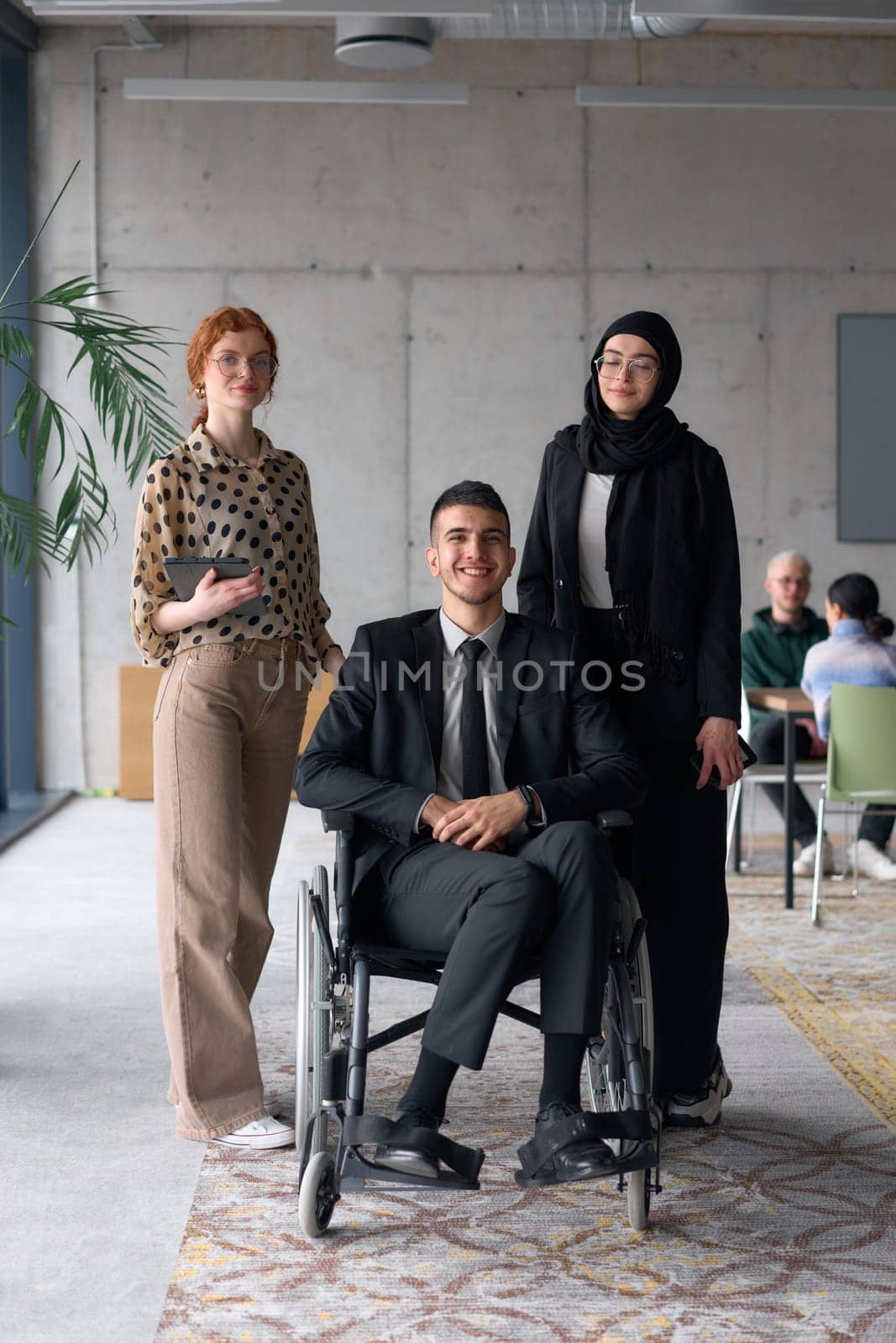 A diverse business team poses confidently, including a smiling director seated in a wheelchair, flanked by two professional female colleagues, one wearing a hijab and the other holding a tablet, in a modern office environment.