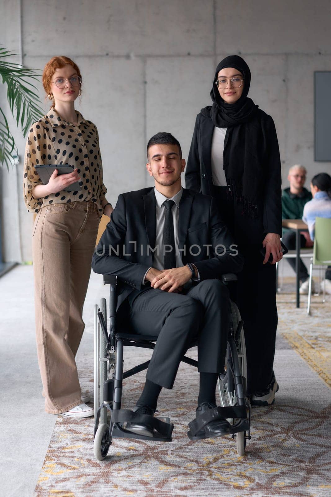 A diverse business team poses confidently, including a smiling director seated in a wheelchair, flanked by two professional female colleagues, one wearing a hijab and the other holding a tablet, in a modern office environment.