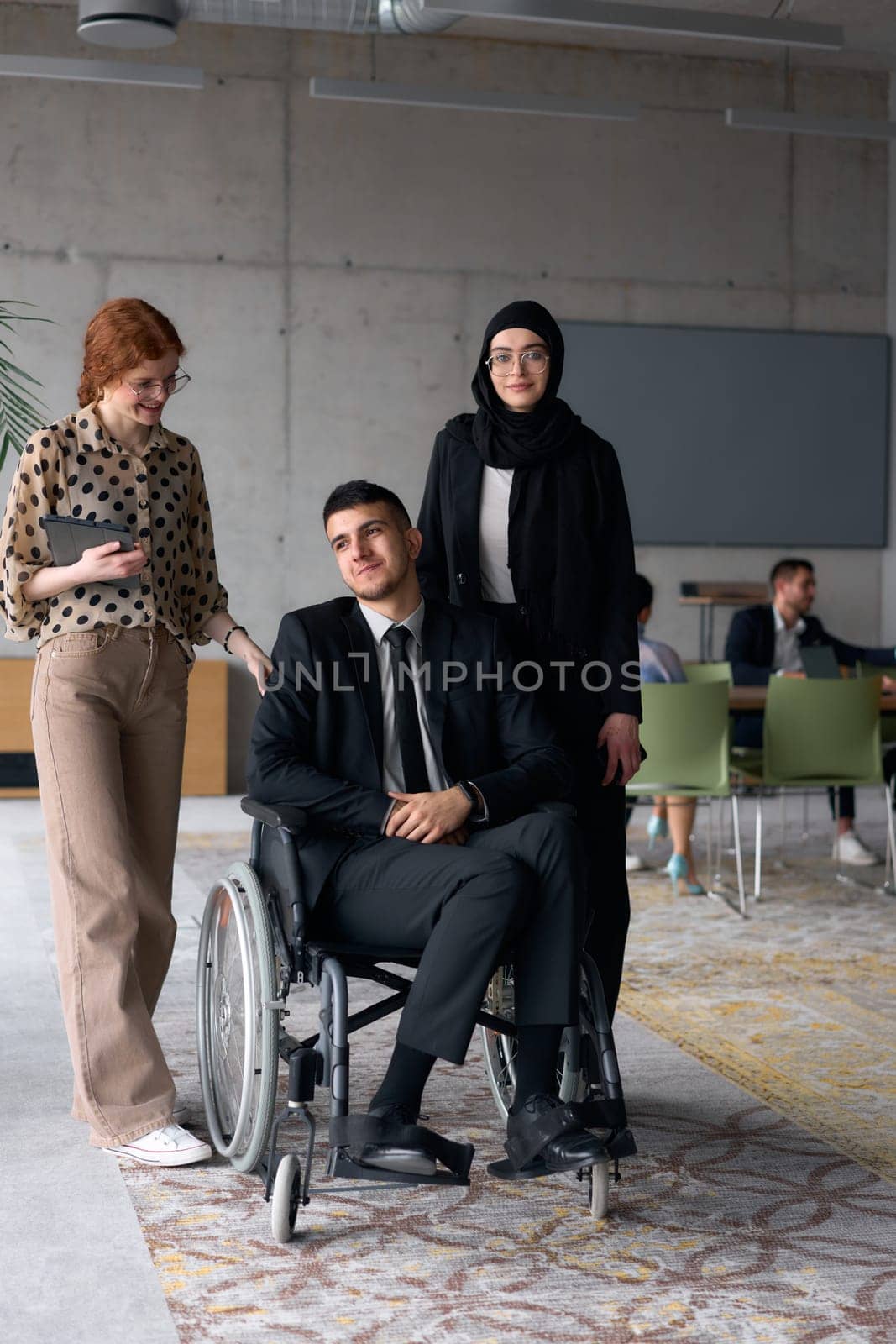 A diverse business team poses confidently, including a smiling director seated in a wheelchair, flanked by two professional female colleagues, one wearing a hijab and the other holding a tablet, in a modern office environment.