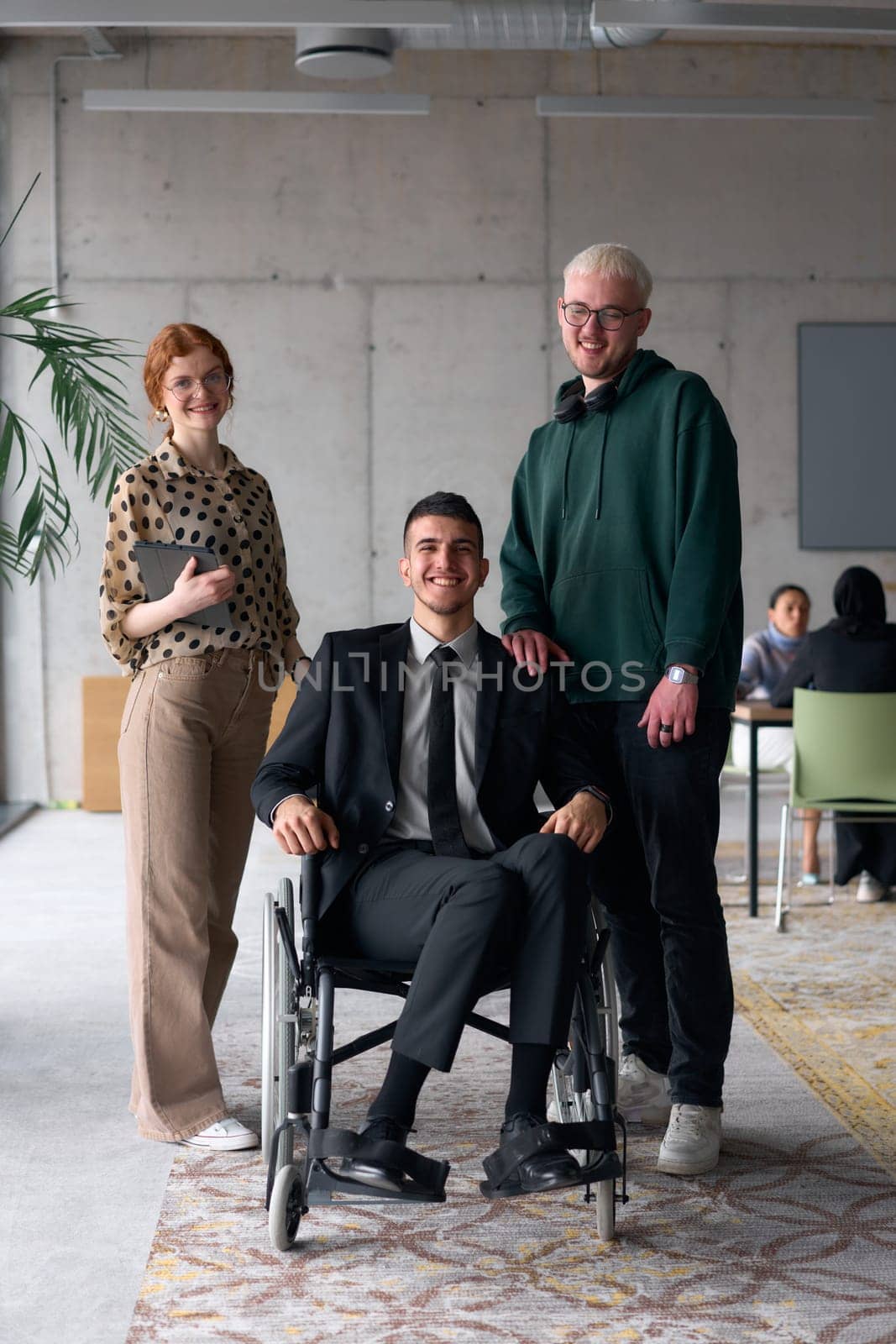 Group cheerful business colleagues, including a businessman in a wheelchair, posing together in a modern office, showcasing teamwork and inclusivity.