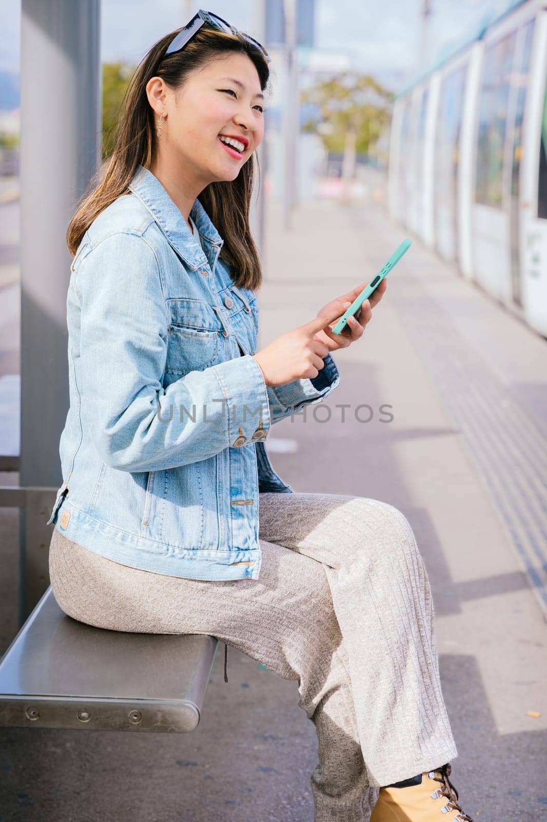 Stylish young woman in a denim jacket sitting at a tram stop, holding her smartphone. She looks to the side with a relaxed expression. Modern urban transportation and city life concept.