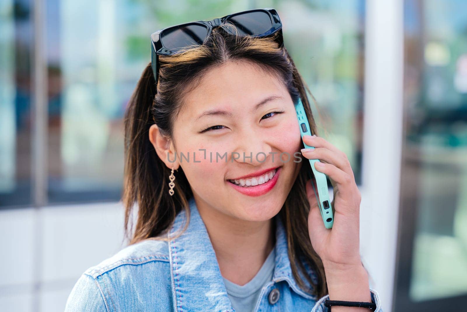 Smiling young woman wearing casual clothes and denim jacket sitting at a tram stop, talking on her mobile phone. Urban setting with tram and city background. Bright and sunny day.