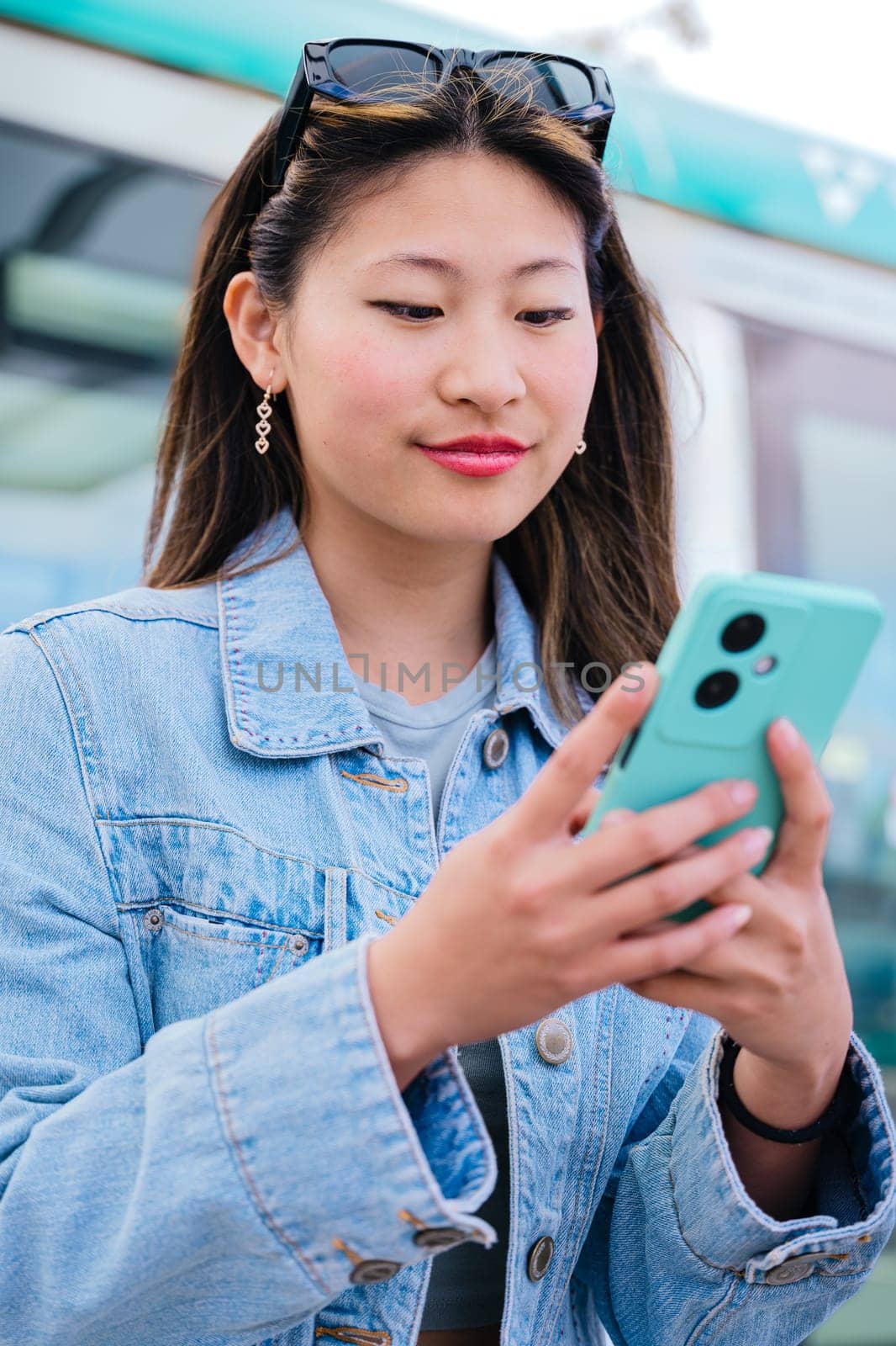 Cheerful young woman in a denim jacket sitting at a tram stop, using her smartphone while waiting for the approaching tram. Modern urban transportation and digital lifestyle concept.Vertical