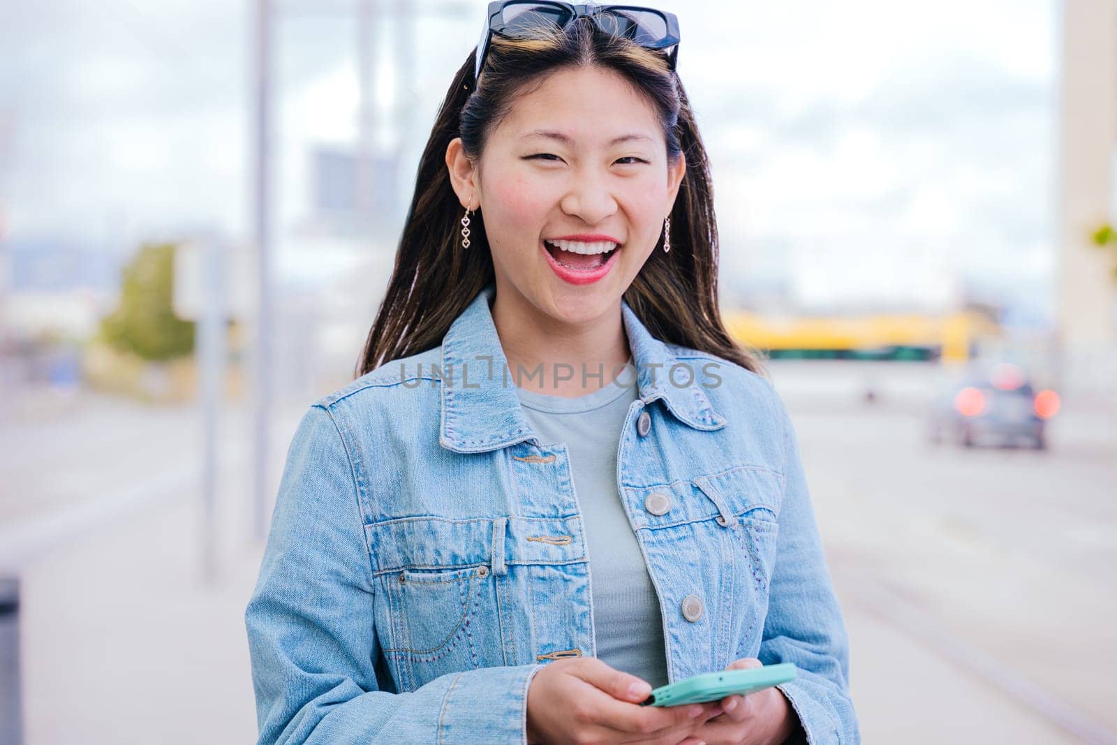 Cheerful young woman in a denim jacket holding her smartphone while looking up with a happy expression. Modern urban setting, representing digital connectivity and city lifestyle.
