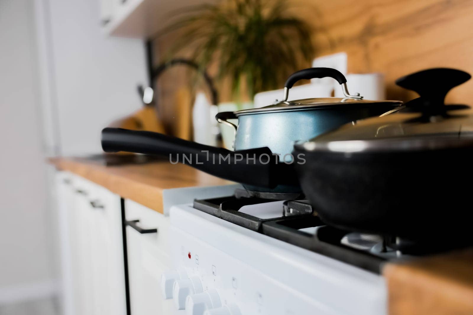 Close-up of a black frying pan with a transparent lid and a silicone handle and a blue saucepan with a transparent lid and silicone handles against the background of a modern wooden-white kitchen out of focus. The concept of cooking, cooking, boiling and frying. High quality photo