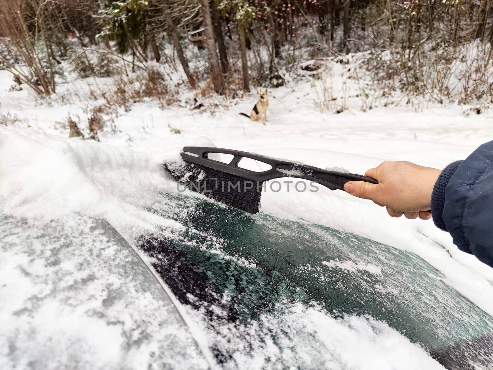 A driver clears snow off their car with a brush as cold winter weather blankets the surroundings in a snowy atmosphere.
