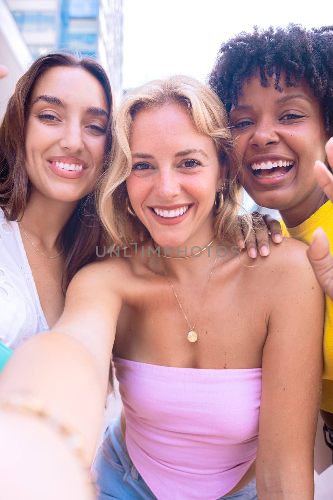 A group of three young women with diverse features smile while taking a selfie in an urban setting. They are dressed in casual summer outfits, enjoying a fun moment outdoors in the city.
