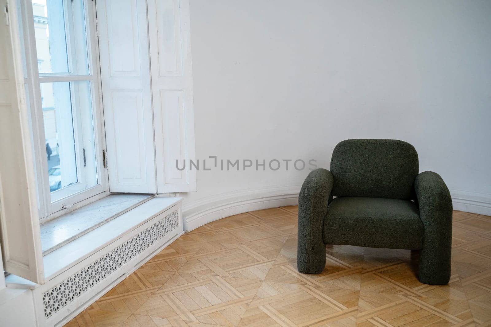 dark gray minimalist style armchair stands in the room. wooden beautiful floor. warm shade parquet lies on the floor. armchair stands against white wall. daylight from large bright window