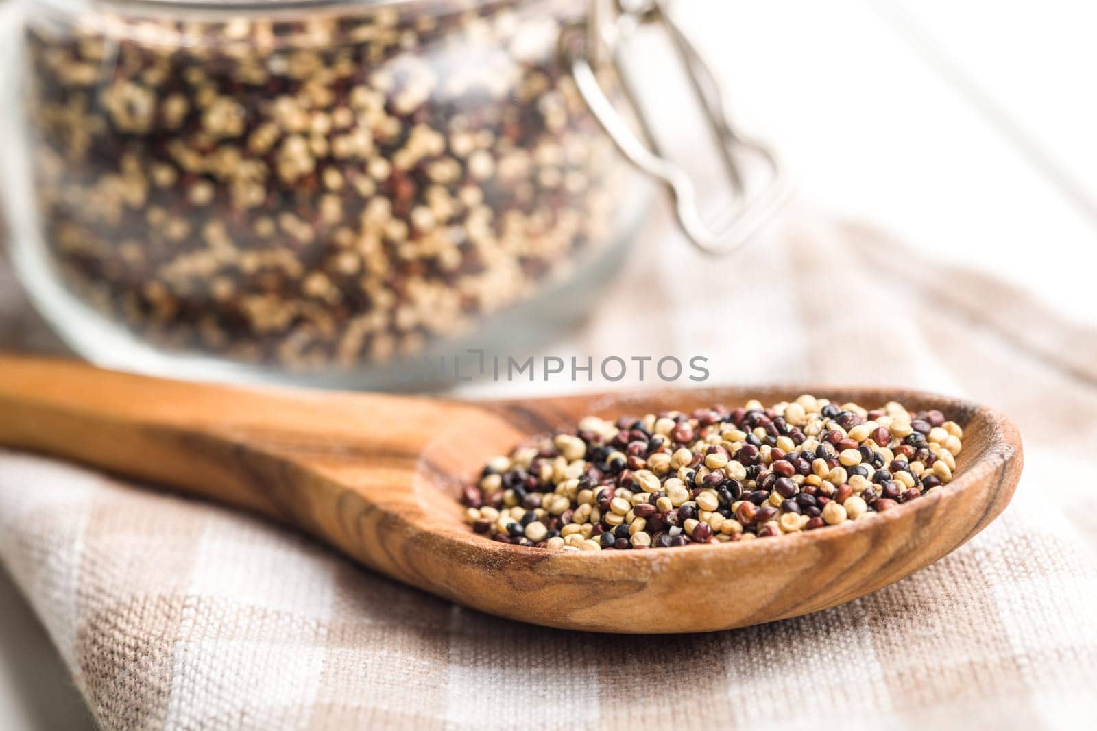 Mixed uncooked quinoa seeds on wooden spoon on a white table.