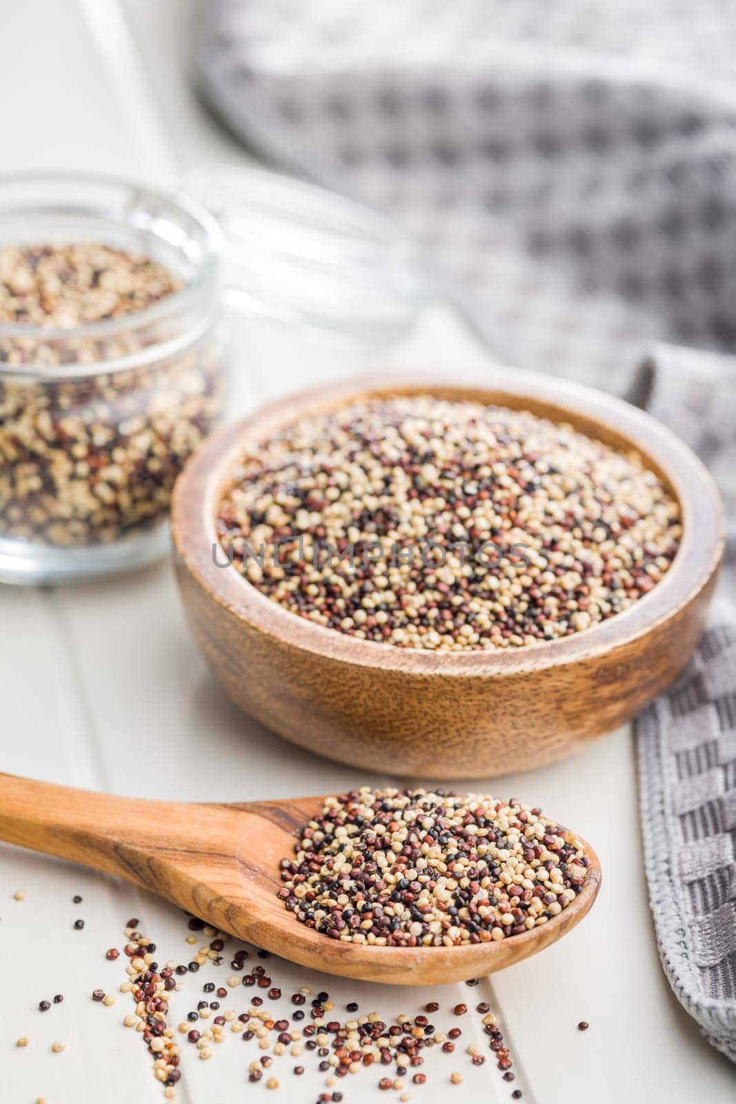 Mixed uncooked quinoa seeds on wooden spoon on a white table.