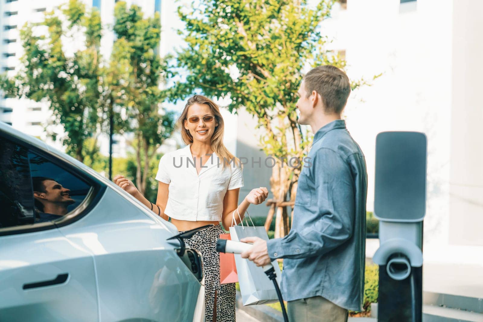 Young couple travel with EV electric car charging in green sustainable city outdoor garden in summer shows urban sustainability lifestyle by green clean rechargeable energy of electric vehicle innards