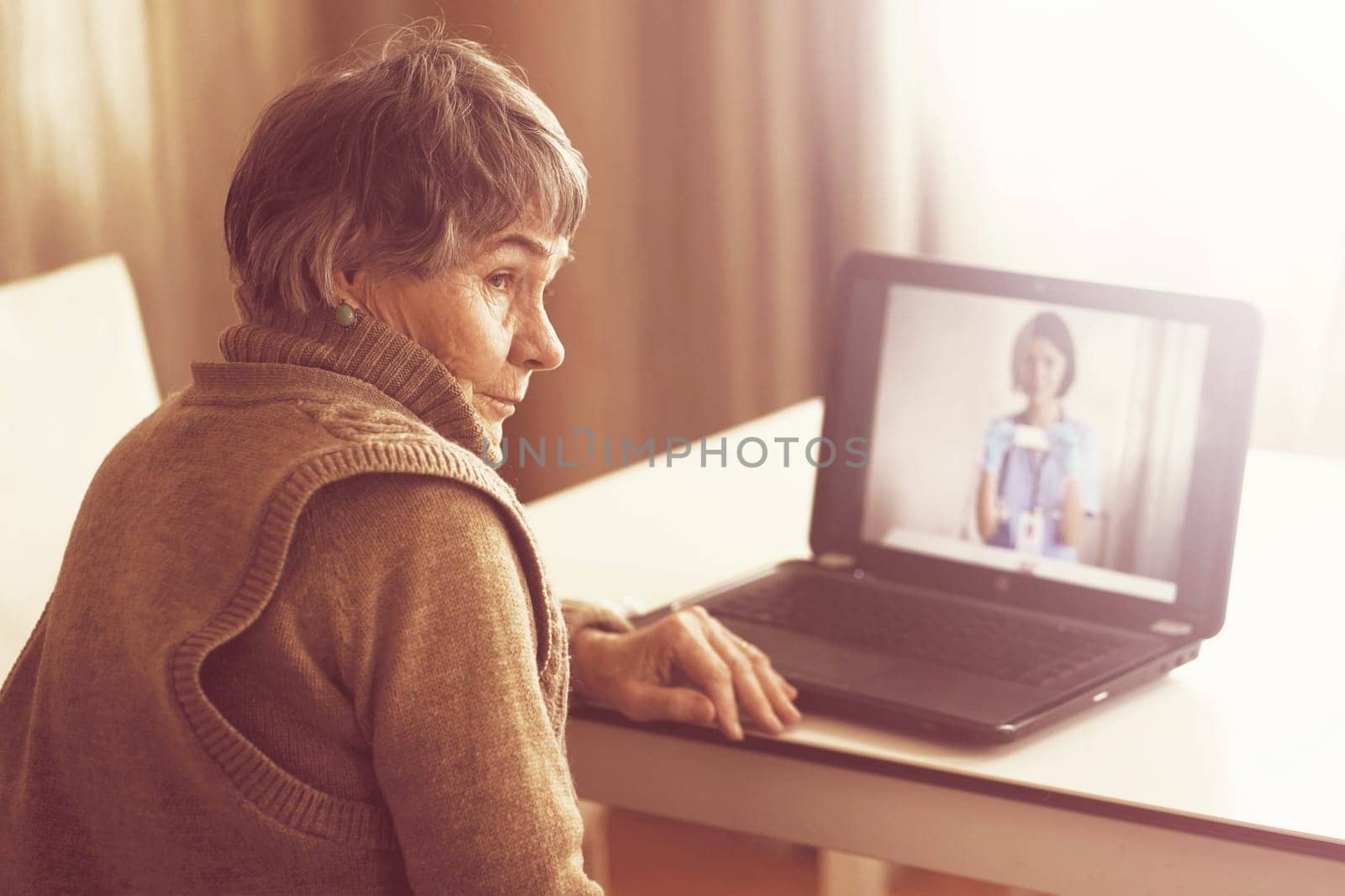 An elderly patient, retired person participates in a digital conversation with a nurse via a webcam. Aged woman talking using a video link with a female doctor, has an online consultation at home.